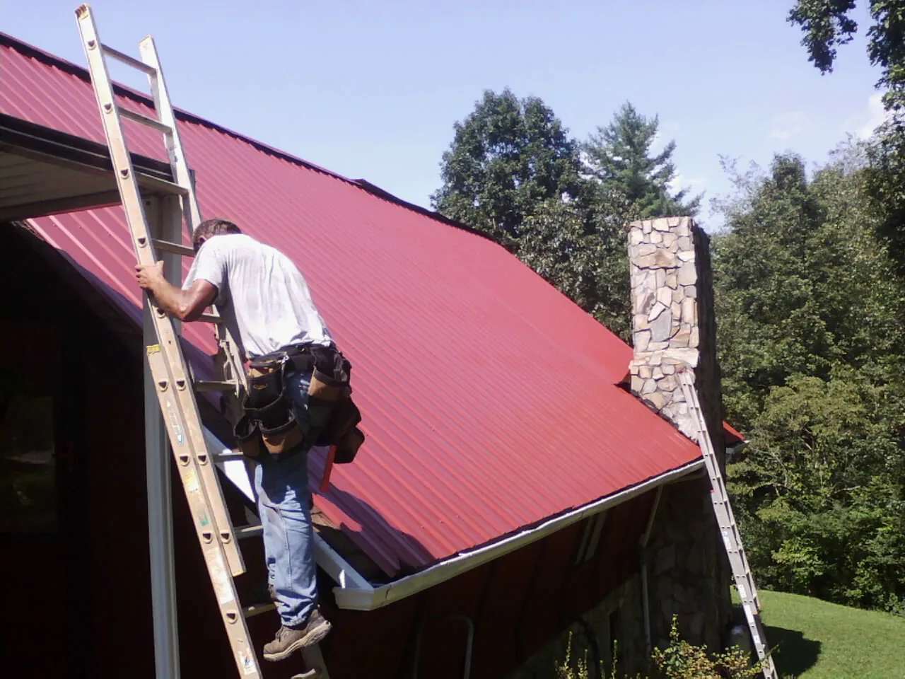 Roofer on a ladder beside a red metal roof and stone chimney — Soffit & Fascia Repair by Roofing Brothers in La Quinta